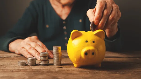 Elderly hands depositing coins into a yellow piggy bank.