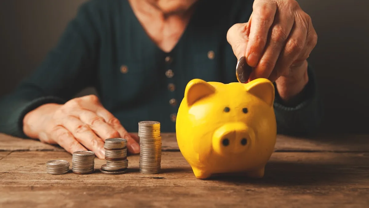 Elderly hands depositing coins into a yellow piggy bank.